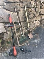 Photo showing garden tools including a battery-powered weed wacker, rakes, shovel, and a tool with a wooden handle against a stone wall