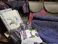Photo showing multiple purple towels, woven black and white place mats, afghans, floral printed fabric, and gray satin-like fabric arranged on and in front of a red bin.