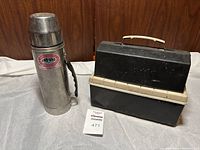 Photo of Unovac stainless steel thermos and black-beige vintage lunch box with auction tag in front