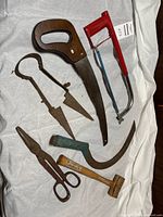 Photo showing all vintage tools laid on white fabric, including wooden hand saw, red hacksaw, green-handled pruning saw, wooden mallet, rusty tin snips, and metal shears.