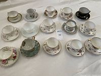 Full overview of 15 teacup and saucer sets arranged on table showing variety in colors, styles, and designs.