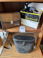 Two carrying cases visible on wooden shelf — brown leather-like case and black hard plastic case alongside the box with Bushnell branding.