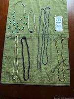 Photo of seven costume necklaces arranged on a green cloth. Included are an Avon faux gold rope chain, two blue beaded necklaces, faux freshwater pearl necklace with gold beads, Chico's triple strand faux turquoise necklace, and black bead strand.