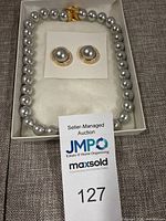 Boxed set of a faux pearl necklace and matching clip-on earrings, showing the necklace with silver round faux pearls, decorated with a gold-tone clasp and matching gold-tone bordered round pearl clip-on earrings.