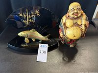 Happy Buddha ceramic figurine and gold tone metal carp on display shown together on a surface.