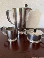 Three-piece stainless steel serving set: coffee/tea pot, creamer, and sugar bowl with lid.