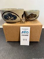 Two Japanese nesting bowls on cardboard box, showing exterior with hand-painted blue and brown floral/leaf motifs and light cream speckled glaze.