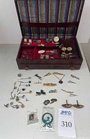 Open jewelry box displaying a variety of cufflinks, tie clips, and pins arranged on red velvet lining and outside of box