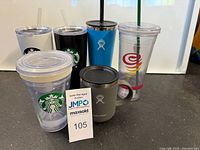 Seven tumblers shown including Starbucks (white and black), Hydro Flask (blue and gray), and two clear plastic tumblers including one with colorful logo and one damaged lid