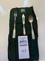 Three-piece flatware set with mother of pearl handles laid out on green felt pouch marked 'Gump's' along with MaxSold auction tag.