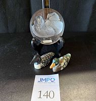 Full view of the Mats Jonasson crystal duck paperweight on wooden stand along with two small wooden duck figurines on a black surface, showing scale and arrangement.