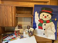 Wide display of lot items including both snowman garden flags, boxed and unboxed Hallmark Keepsake ornaments, multiple snowman sculptures on table beneath wooden cabinet