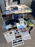 Wide view of all items arranged on tables including five etched glasses, prints, brochures, envelopes, coins, stamps, pen and ephemera