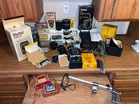 Photo showing a large selection of vintage camera equipment laid out on a kitchen counter including Brownie Reflex camera, lenses, flashes, accessories.