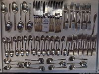 Full view of the flatware set showing knives, forks, spoons, teaspoons, serving spoons, and butter spreader arranged in a display case with a label.