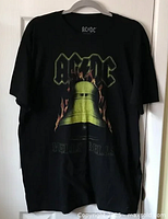 Black unisex T-shirt hanging on hanger with front full-color graphic showing AC/DC logo and flaming bell