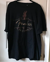 Front view of the black Fender T-shirt hanging against a white door, showing the full design and shirt shape.
