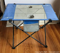Front angle of the blue folding camping table set up on a wooden floor, showing the mesh top with two circular mug holders and a side mesh pocket below.