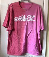 Full front view of the pink two tone Gorillaz t-shirt on a hanger showing the large white brush font band logo.