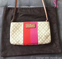 Front view of Kate Spade crossbody bag showing beige and cream monogram jacquard fabric with red and pink vertical stripes and gold-tone brand nameplate. Caramel leather shoulder strap and edges visible. Bag placed on dark brown dustbag.