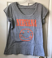 Full view of the light grey Nirvana t-shirt hanging on a door, showing the orange Nirvana logo and smiley face graphic on the front