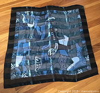 Full scarf laid flat on wooden floor showing overall abstract design with black border, blue, gray, and white colors in geometric shapes including a face and various prints.