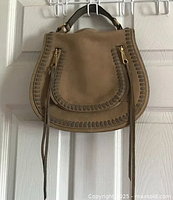Front view of taupe suede saddle bag showing leather whipstitch detailing and gold-tone zipper pulls with tassels
