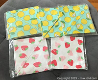 Photo of the 5 cloth napkins sealed in plastic with visible lemon and strawberry prints, arranged on a dark grey surface.