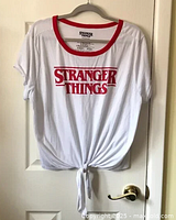 Front view of white tie front t-shirt with red Stranger Things logo and red collar hanging on door