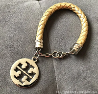 Gold woven leather rope bracelet with braided texture, silver toned end caps, lobster claw clasp, and large silver toned Tory Burch logo charm.