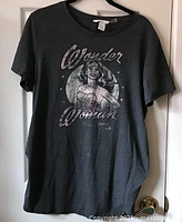 Full view of a grey Wonder Woman t-shirt hanging on a door, showing overall garment including sleeves and neck.