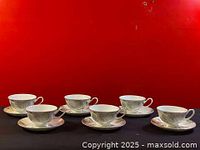 Six bone china teacups with matching saucers against a red background, showing front view of cups and saucers on a table.