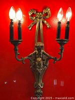 Full view of brass Louis XVI style two-armed wall sconce illuminated with 4 bulbs against a red background showing detailed bow and cherub motifs.