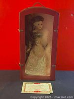 Porcelain doll in enclosed glass-front showcase, dressed in gold embroidered gown with feather boa, against red background.
