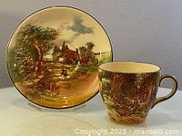 Cup and saucer together showing matching landscape scenes in brownish/green tones on cream porcelain.