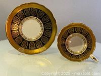 Teacup and saucer set showing black and gold gilded leaf floral decorative pattern with scalloped edges, items displayed on clear stands