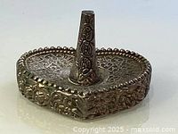 Close-up of the silver heart-shaped ring holder showing detailed floral and scroll engravings on the base and cone stand.
