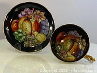 Front view of cup and saucer showing colorful fruit painting and gold accent on handle against black background.
