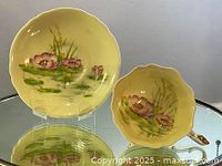 Photo showing the tea cup with scalloped edges and hand painted water lily flowers in pink and yellow with green lily pads on a yellow background, alongside the matching saucer with similar design.