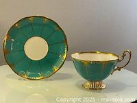 Front view of turquoise and gold teacup beside matching saucer on a white surface