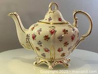 Side view of the teapot showing the curved spout, floral pattern, and gold accents on the rim and handle.