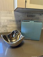 Polished Nambe Wave Bowl next to original blue box on kitchen countertop, showing side view and wave-like rim design.