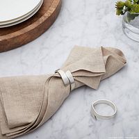 Close-up of one brushed stainless steel napkin ring holding a beige cloth napkin on a marble surface next to plates and a small flower vase.