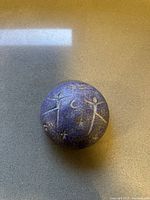 Top side of the round porcelain art ball showing blue-violet glaze and carved primitive stick figures with stars and moon
