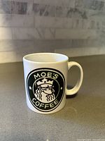 Photo of a white ceramic coffee mug featuring a black and white Moe's Coffee logo inspired by Starbucks, placed on a gray countertop with a tiled backsplash.