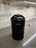 Front view of black cylindrical ToLife air purifier on countertop, showing top vent and brand name.