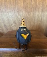 Front view of vintage black pottery crow decoration holding a candy corn piece in its beak, wearing an orange pointed hat with black polka dots.