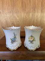 Front view of two white porcelain vases with floral hand painted designs and gold accented scalloped rims and base.