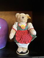 Front view of vintage Steiff mohair teddy bear wearing green and red dress on stand, showing Original Steiff tag on chest and yellow Steiff ear tag.