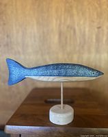 Side view of the hand painted wooden fish sculpture showing the blue speckled upper body, white underside, yellow eye, and wooden base.